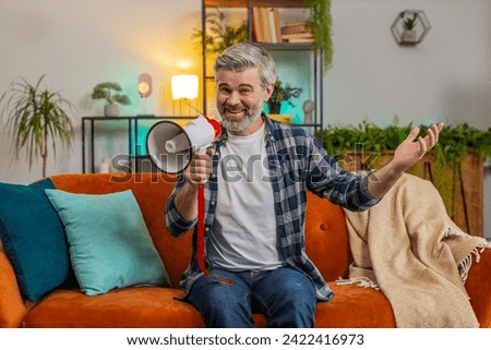 Happy senior man talking into megaphone, proclaiming news, loudly announcing advertisement, warning using loudspeaker to shout speech. Caucasian mature guy announcing big sale sitting on sofa at home