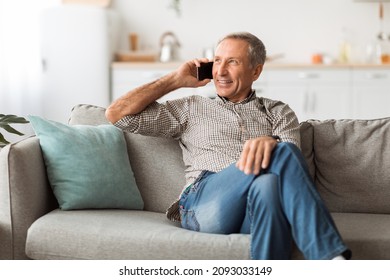 Happy Senior Man Talking On Mobile Phone Looking Aside Sitting On Couch At Home. Male Communicating Having Cellphone Conversation Indoor. Modern Communication, People And Gadgets Concept