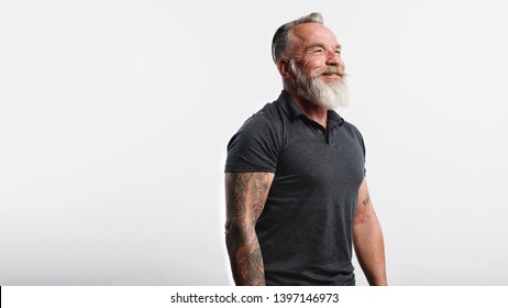 Happy Senior Man With Tattoo On Arms Standing Against White Background. Portrait Of Old Muscular Male With A White Beard Looking Away.