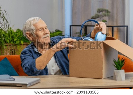 Happy senior man unpacking delivery parcel with headphones at home. Smiling Caucasian grandfather shopper, online shop customer opening cardboard box receiving purchase gift by fast postal shipping
