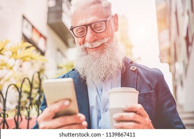 Happy senior man using smartphone app outdoor while drinking take away coffee - Fashion old guy having fun with new trend technology - Tech and joyful elderly lifestyle concept - Focus on his mout