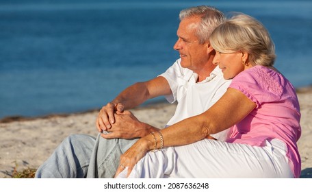 Happy Senior Man And Woman Retired Couple Sitting, Smiling On A Sunny Beach 