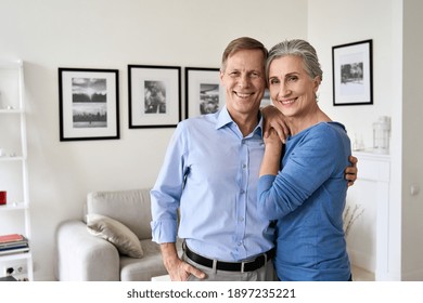 Happy Senior Mature 60s Family Couple Hugging, Looking At Camera, Standing In Living Room In Modern Apartment. Smiling Satisfied Middle Aged Husband And Wife Embracing Posing For Portrait At New Home.