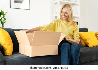 Happy Senior Mature Adult Woman Is Looking At Her New Parcels, Sitting On The Couch, And Unpacking. Smiling Lady Opening Online Store Order In The Postal Delivery Shipping Box