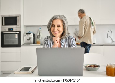 Happy Senior Mature Woman Wife Remote Working Or Distance Learning Online From Home Office Using Laptop Computer While Her Old Husband Cooking Breakfast In Kitchen. Middle Aged Family Couple At Home.