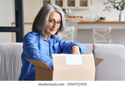 Happy senior middle 60s aged woman opening box with ordered clothes at home on couch. Old mature online shopper customer opening online shop parcel. International delivery service comfort concept.
