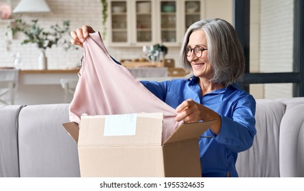 Happy Senior Middle 60s Aged Woman Opening Box With Ordered Clothes At Home On Couch. Old Mature Online Shopper Customer Smiling And Holding Dress. Delivery Service Fashion Comfort Concept.