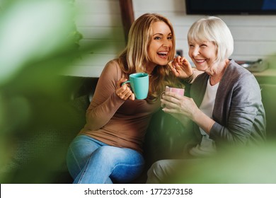 Happy Senior Mother With Adult Daughter Sitting On Couch And Holding Cups With Coffee Or Tea At Home. Togetherness Concept