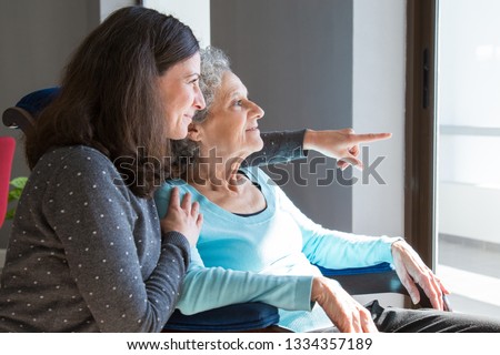 Happy senior mother and adult daughter enjoying dramatic view out of window. Elderly lady resting in armchair, while young woman pointing out of window. Scene from window concept