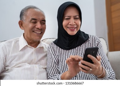 Happy Senior Muslim Couple Using Mobile Phone In Living Room. Old Asian People