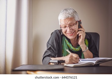 Happy Senior Old Business Woman Writing Down Orders, Talking On Her Cell Phone