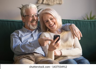 Happy Senior Old Couple Holding Smartphone Looking At Cellphone Screen Laughing Relaxing Sit On Sofa Together, Smiling Elder Mature Grandparents Family Embracing Having Fun With Mobile Phone At Home