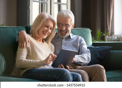 Happy Senior Old Couple Laughing Relax Sit On Sofa Using Digital Tablet Make Distance Video Call Doing Internet Shopping Together, Elder Mature Grandparents Embrace Looking At Computer Screen At Home