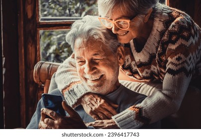 Happy senior old couple use mobile phone at home together to video call parents. Winter season indoor leisure activity for retired mature people. Woman hug mature man on the chair. Love and technology - Shutterstock ID 2396417743