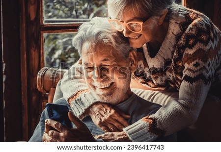 Happy senior old couple use mobile phone at home together to video call parents. Winter season indoor leisure activity for retired mature people. Woman hug mature man on the chair. Love and technology