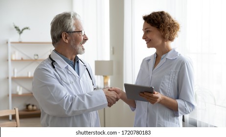 Happy Senior Old Male Physician Handshaking Young Female Nurse Thanking For Help In Professional Teamwork. Two Doctors Team Shake Hands Expressing Gratitude, Trust In Medical Collaboration Concept.