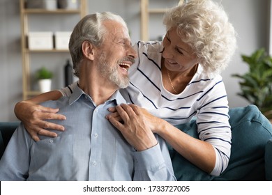 Happy Senior Old Woman Embracing From Back Joyful Mature Husband, Sitting On Comfortable Sofa. Emotional Positive Middle Aged Family Couple Enjoying Pleasant Conversation Talking Chatting At Home.