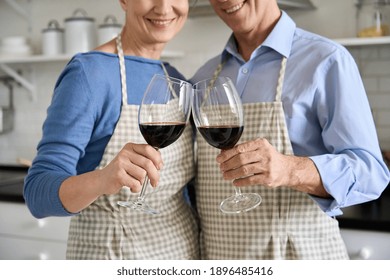 Happy Senior Older Couple Husband And Wife Wearing Aprons Holding Clinking Glasses In Hands, Drinking Red Wine, Toasting, Standing In Kitchen Together. Wineglasses Glassware Concept, Close Up View.