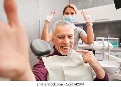 Happy Senior Patient And His Female Dentist Taking Selfie Together. Patient Is Holding Smartphone And Showing To His Healthy Smile With His Finger. Stock Photo