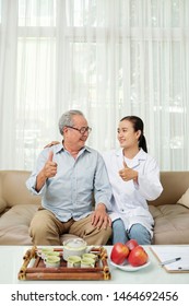 Happy Senior Patient And Young Nurse Showing Thumbs Up And Looking At Each Other While Sitting On Sofa And Drinking Tea Nurse Coming To Visit Him At Home