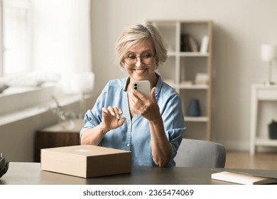 Happy senior pensioner lady using online post office service on mobile phone, sitting at table with received parcel, carton box from delivery service, giving feedback to Internet store - Powered by Shutterstock