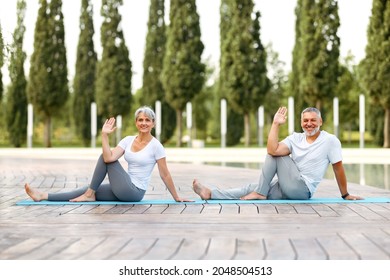 Happy Senior Retired Couple Doing Partner Yoga Outside In City Park, Mature Active Husband And Wife Sitting Barefoot On Mats In Eka Pada Upavistha Parivrttasana Pose Looking At Camera With Smile