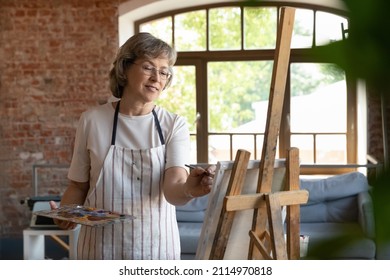 Happy senior retired lady drawing in paints on canvas. Elder talented professional artist working at easel in artistic studio, using palette, paintbrush, smiling, enjoying creativity, inspiration