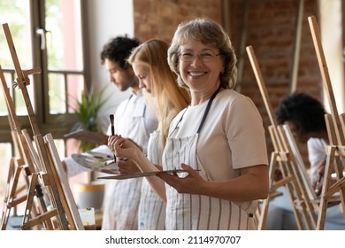 Happy senior retired student of art school drawing in class, using artist tools, canvas, standing at easel, looking at camera, smiling, laughing. Head shot portrait in artistic studio