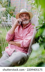 Happy Senior Sitting In Outdoor Garden Swing And Phoneing.