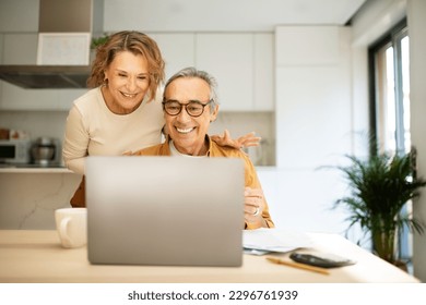 Happy senior spouses paying bills online from home, sitting in front of laptop with papers, enjoying easy Internet banking, kitchen interior, copy space - Powered by Shutterstock