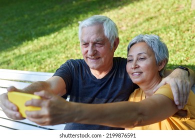 Happy Senior Spouses Take Selfie Picture Sit On Bench In Summer Park. Grey-haired Couple Look At Cell Phone Screen Make Videocall Outdoor Use Modern Tech, Enjoy Online Fun Together Spend Time Outside