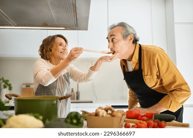 Happy senior spouses tasting food, woman giving her husband to try soup, cooking healthy lunch together in kitchen interior, man helping wife preparing meal - Powered by Shutterstock