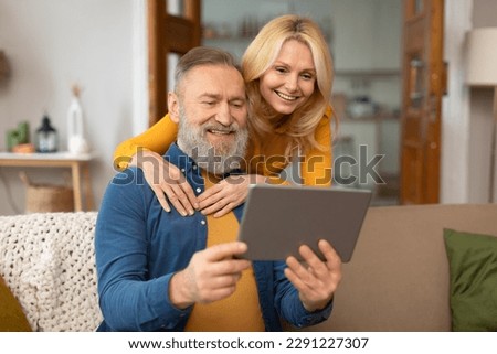 Happy Senior Spouses Using Digital Tablet Computer Browsing Internet And Watching Movie Online Sitting On Couch In Modern Living Room At Home. Internet And Technology Concept