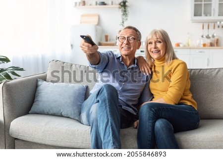 Happy Senior Spouses Watching TV Pointing Television Remote Control Sitting And Hugging On Sofa At Home. Mature Couple Enjoying Favorite Movie Together On Weekend. Retired Family Leisure