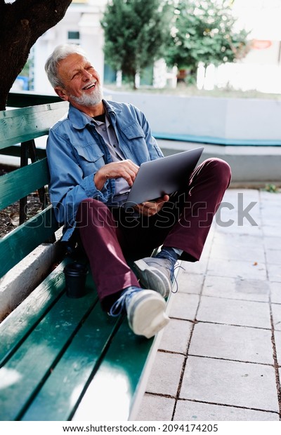 A happy senior\
student sits on the bench in a park and holds a laptop in his lap.\
Senior students in\
retraining.