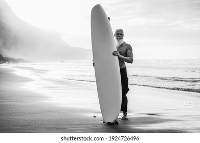 Happy Senior Surfer Holding Surf Board On The Beach At Sunset - Focus On Face