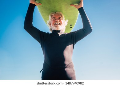 Happy Senior With Surftable On His Head Is Smiling And Laughing - Old And Mature Man Having Fun Surfing With A Black Wetsuits - Active Retired Adult Doing Activity Alone