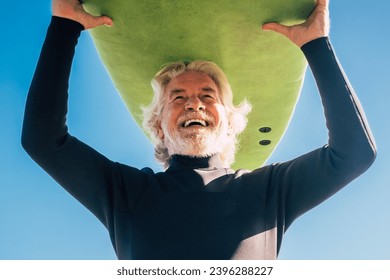 happy senior with surftable on his head is smiling and laughing - old and mature man having fun surfing with a black wetsuits - active retired adult doing activity alone - Powered by Shutterstock