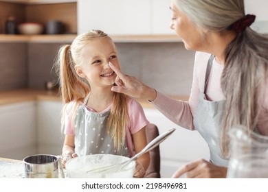 Happy Senior Woman In Apron Touches Nose Of Little Girl, Make Dough And Have Fun With Flour, Family Enjoy Leisure Time In Kitchen Interior. Cooking Lesson Baking And Prepare Homemade Food Together