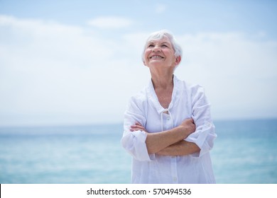 Happy senior woman with arms crossed looking up while standing at sea shore - Powered by Shutterstock