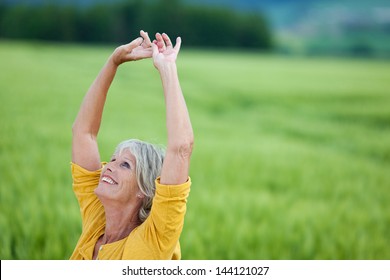 Happy Senior Woman With Arms Raised Looking Up While Standing On Grassy Field