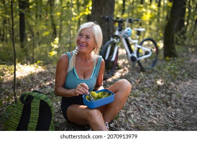 Happy Senior Woman Biker Resting, Sitting And Eating Snack Outdoors In Forest.