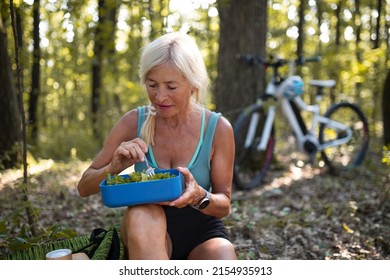 Happy Senior Woman Biker Resting, Sitting And Eating Snack Outdoors In Forest.