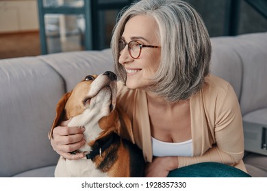 Happy senior woman in casual clothing spending time with her dog while sitting on the sofa at home