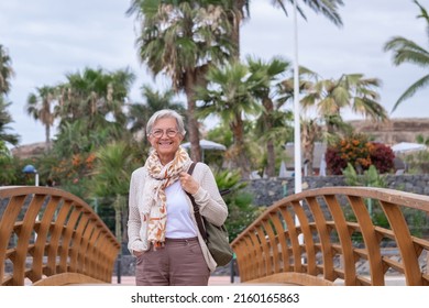 Happy senior woman casual dressed walking on a wooden bridge smiling looking at camera. Palm trees on background - Shutterstock ID 2160165863