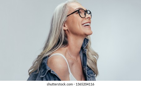 Happy Senior Woman In Casuals Looking Away And Smiling Over Grey Background. Cheerful Mature Woman In Denim Shirt And Eyeglasses.