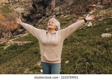 Happy senior woman celebrating with her arms outstretched while standing on a hill. Cheerful senior woman enjoying a leisurely hike outdoors. Woman enjoying recreational activities after retirement. - Powered by Shutterstock