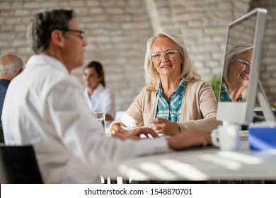 Happy Senior Woman Communicating With Health Care Provider While He Is Analyzing Medical Data On A Computer At Clinic. 