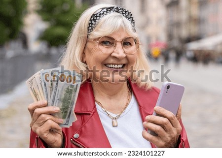 Happy senior woman counting money dollar cash, use smartphone calculator app, satisfied of income for planned vacation gifts. Elderly grandmother walking in urban city street. Town lifestyles outdoors