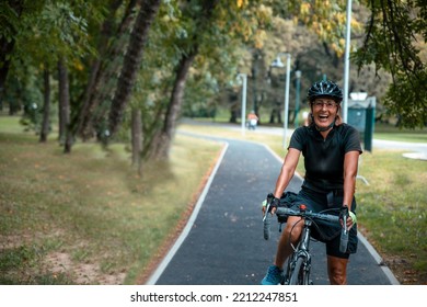 Happy Senior Woman Cyclist Portrait In Black Sportswear And Helmet. Smiling Female Riding Bicycle Outdoor In The City Park. Copy Space